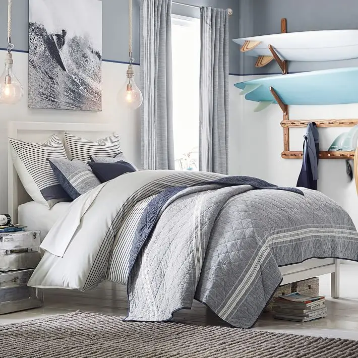 Surfer’s Haven bedroom: Grey and white walls, blue-grey bedding, glass pendants and a rack of surfboards as the feature