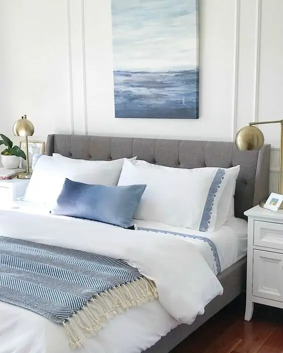 Ocean Hues coastal bedroom: A grey upholstered bed, white sheets with blue trim, brass lamps and one sea painting. A single